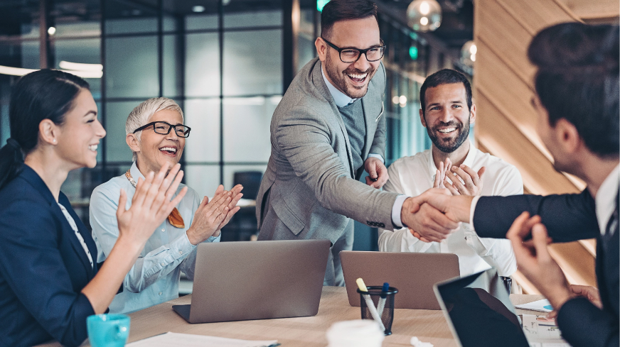 A team of agents celebrates with a client. Two people shake hands across a table while the rest clap their hands